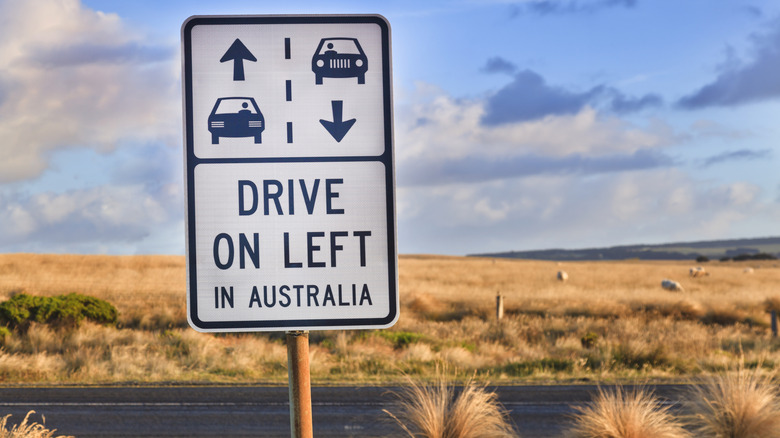 Sign displaying 'Drive On Left In Australia' against a rural road and backdrop.