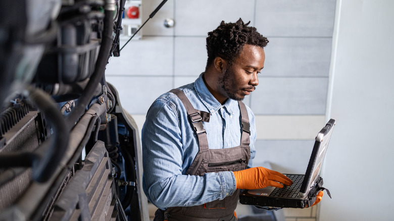 Mechanic in overalls and orange gloves running automotive diagnostics on laptop