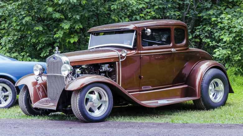 A brown Ford Model A next to a blue 60s muscle car, front 3/4 view