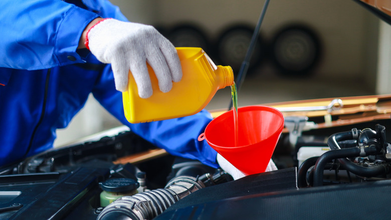 A mechanic wearing yellow gloves pouring engine oil through a red funnel in a car's engine.