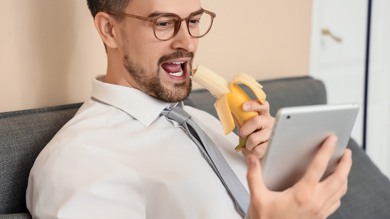 A young businessman sitting on a sofa with a tablet computer eating a banana.