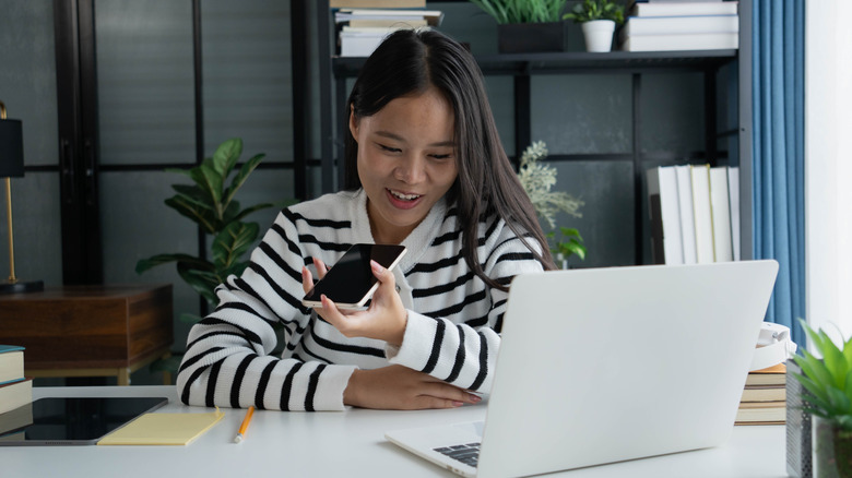 Woman at a desk speaking into a smartphone while looking at a laptop