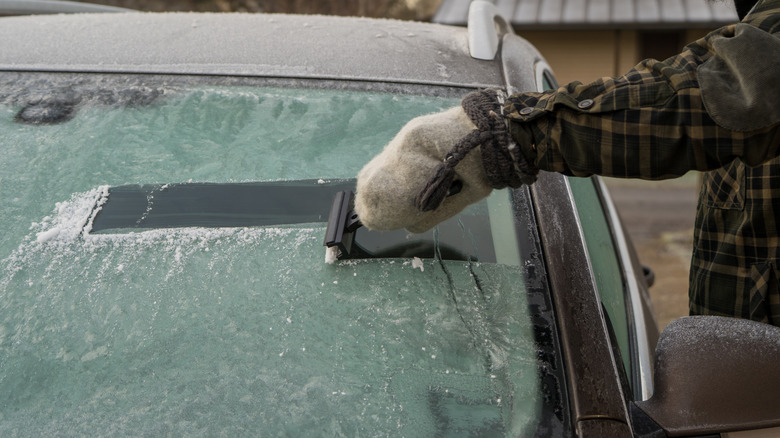 A person in a plaid coat scraping ice from their car's windshield