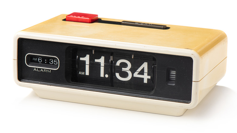 Vintage flip alarm clock. The clock has a cream color which may be discolored white. The clock face is black with white numbers and shows the time is 11.34 a.m. with an alarm set for 6.35 a.m. There is a large red snooze button on top.