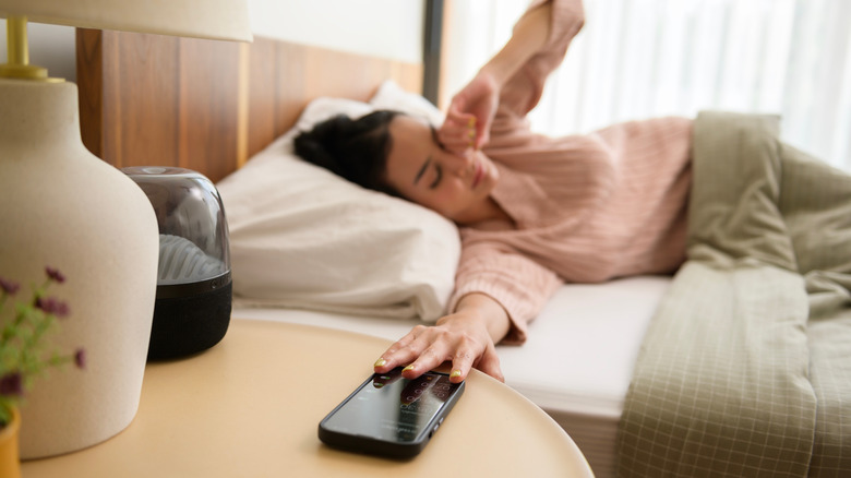 A woman with dark hair wearing pink pyjamas is lying in a bed with green and white covers. She is rubbing sleep from her eyes with her left hand and her right arm is stretched forward towards the smartphone on her bedside table.