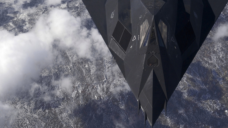 An F-117 Nighthawk flying over clouds and mountains