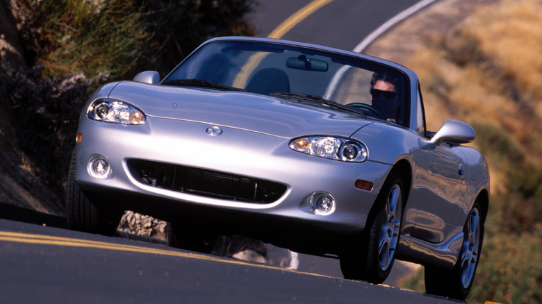 Front 3/4 view of man driving silver Mazda Miata with top down on winding road