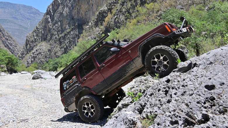 A maroon Jeep Cherokee XJ with large, aftermarket wheels at a steep upward angle in a rocky gorge.