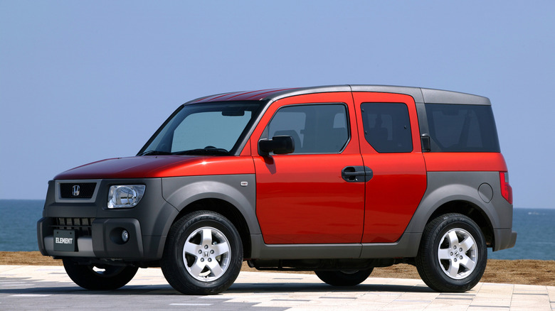 A red Honda Element parked next to a coast, front 3/4 view