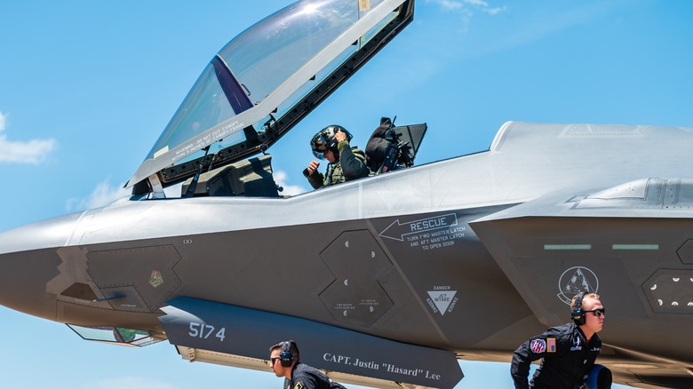 A pilot sits inside the cockpit of an F-35 while ground crew personnel run beside the aircraft