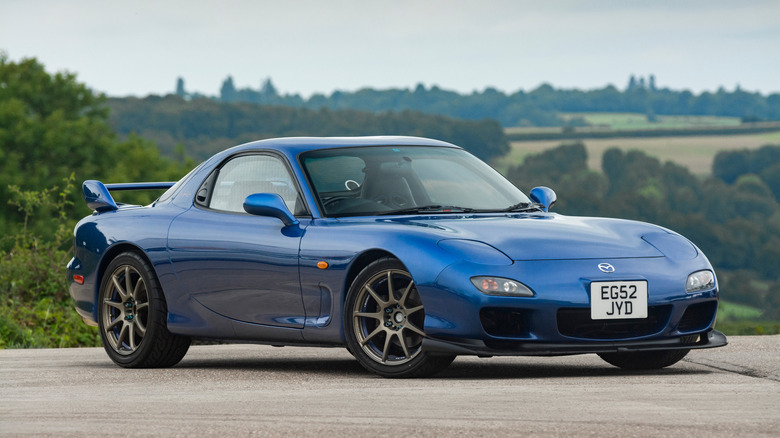 Front 3/4 view of the FD3S Mazda RX-7 in a deep blue paint with a countryside behind it