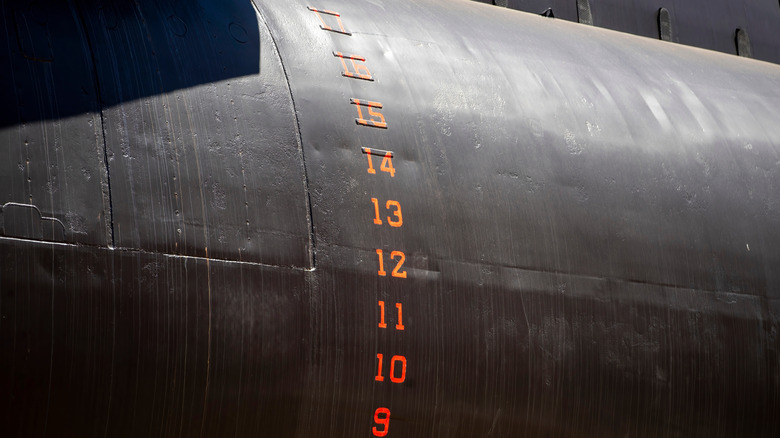 Close-up of a submarine's hull showing depth markings in red, with visible surface textures and light reflecting off the dark metal.