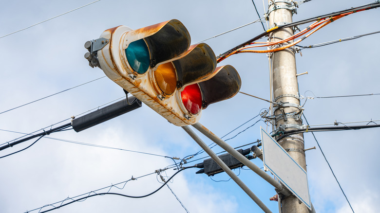 A rusted traffic signal showing blue, amber, and red lights, plus a cloudly sky and electrical wires behind it.