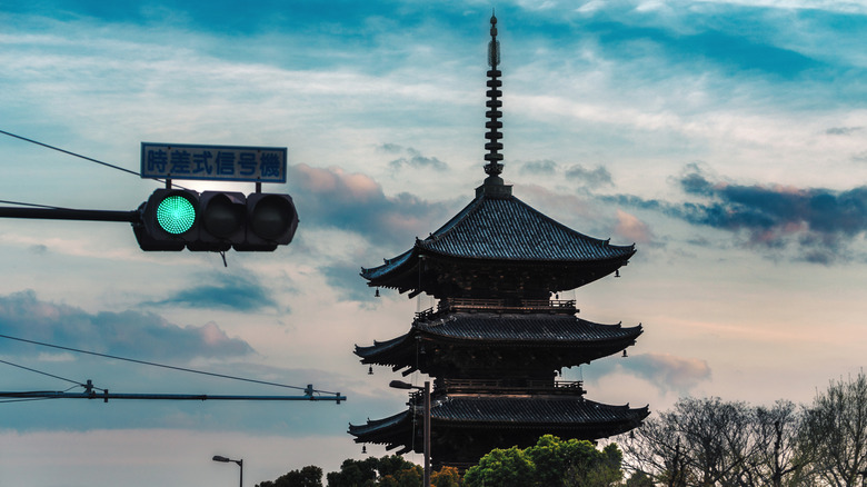 A traffic signal lit up green with two dim lights on a cloudy sky with one of Kyoto's temples behind it. A sign above the light says time-operated traffic light.