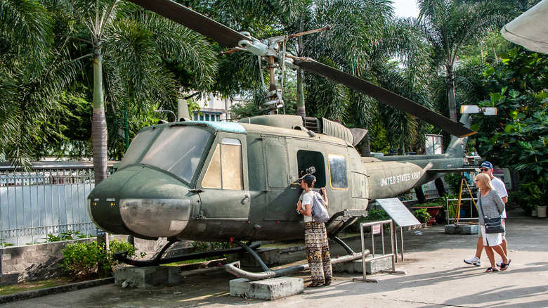 People walking by looking at an old Huey helicopter on cement blocks