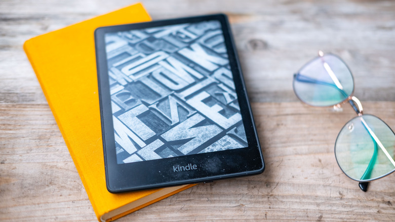 A Kindle e-reader shown sitting on a hardcover next to a pair of glasses