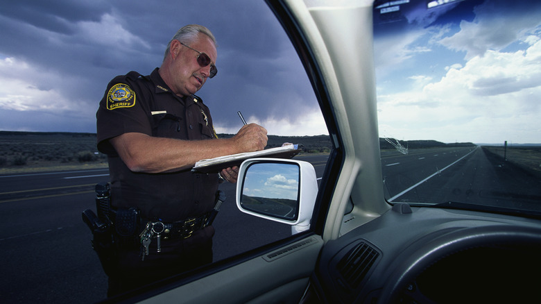 Sheriff standing outside a car writing on a clipboard. The wing mirror and interior of car are visible.