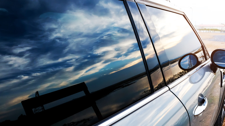 Dark tinted film on siver car's windows reflecting the sky, beach and a wooden bench.