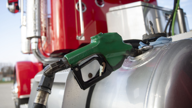 Close up view of an unattended, green fuel nozzle in the fuel tank of a red semi-truck tractor.