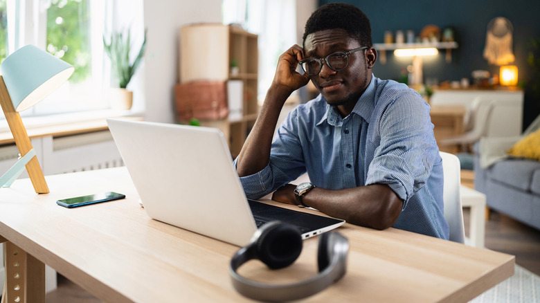 person frowning at laptop screen with unused headphones sitting on table in foreground