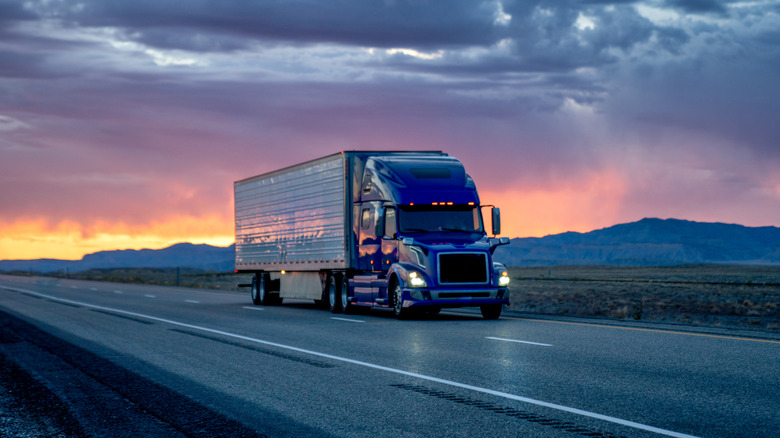 A commercial truck on an empty road at dawn with its headlights lit