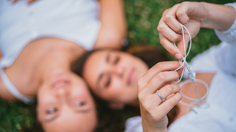 Two people lying in the grass, one holding white wired Apple headphones up to the camera to untangle them.