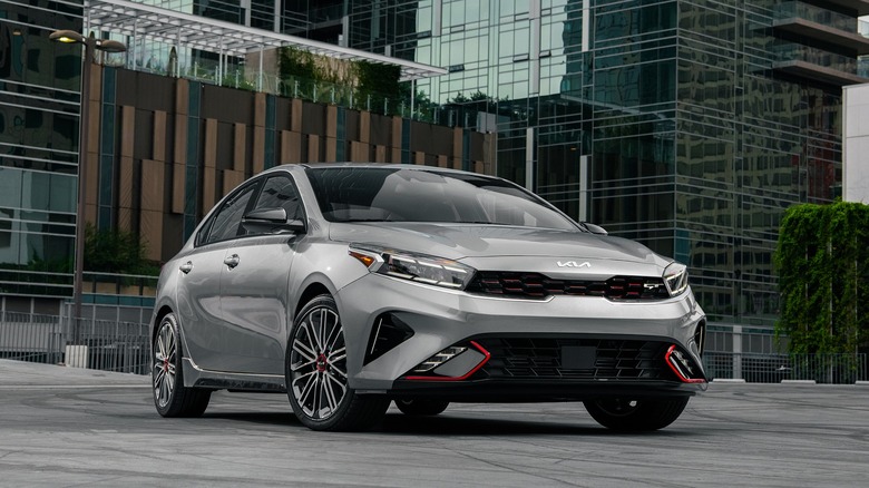 A view of a 2024 Kia Forte sedan in silver, parked in an urban setting with modern glass buildings in the background.