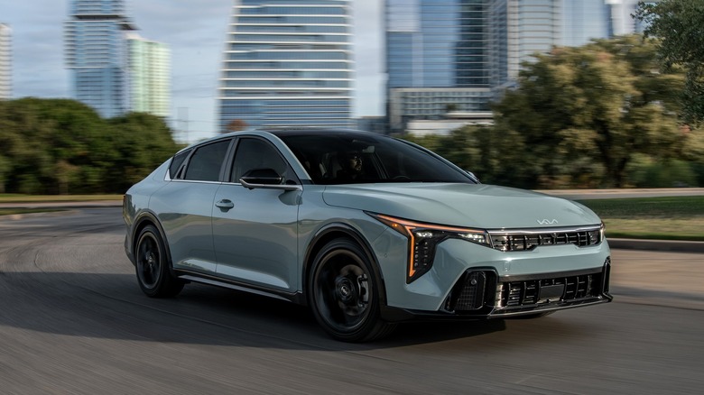 A 2025 Kia K4 in motion on a city road, with modern high-rise buildings and trees blurred in the background.