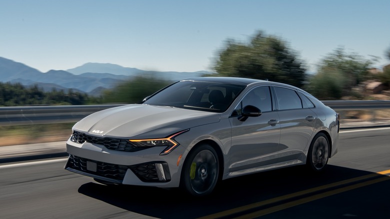 A silver 2025 Kia K5 driving along a scenic mountain road.