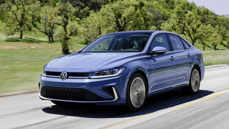 A blue 2025 Volkswagen Jetta driving on a scenic road surrounded by green hills and trees.