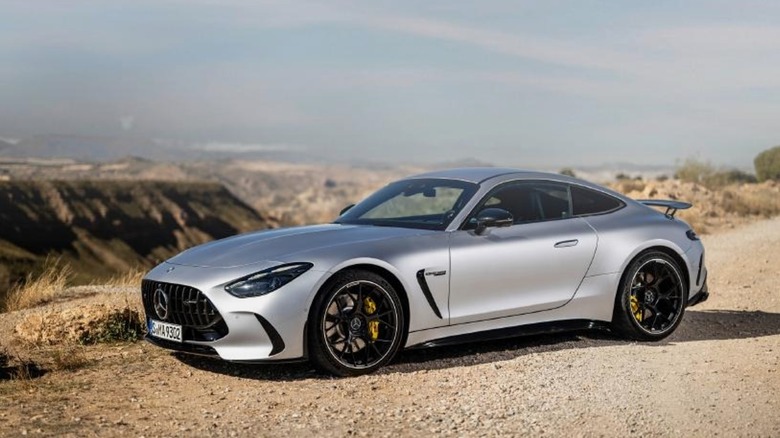 A silver Mercedes-AMG coupe parked on a dirt road with a scenic mountain backdrop.