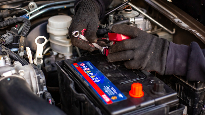 A man installs a battery by tightening the terminals with a wrench under the hood of a car.