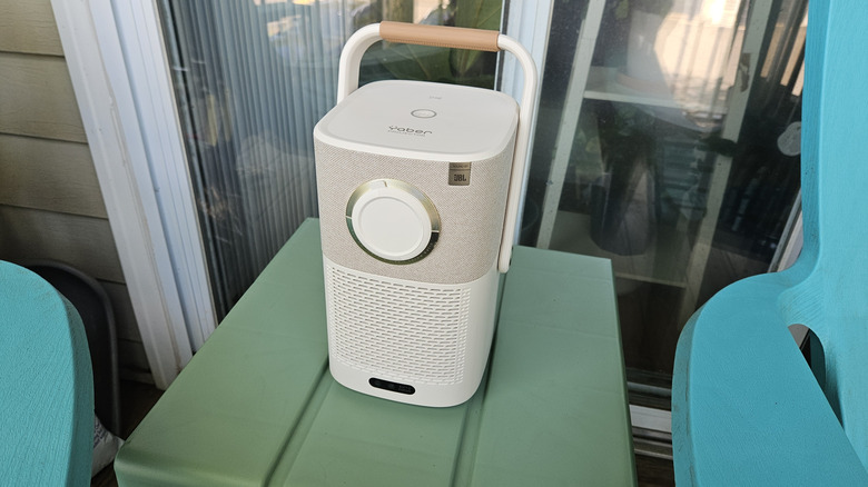 The Yaber T2 Plus Outdoor Projector on a green table with its handle up and lens cap on.