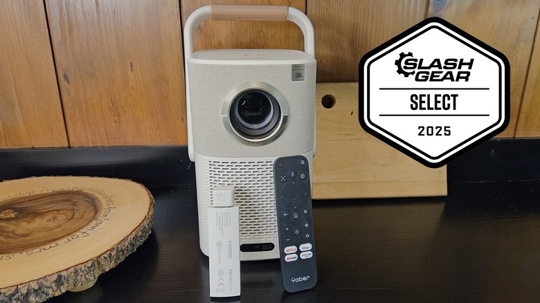 The Yaber T2 Plus Outdoor Projector on a black shelf with a wood background with its remote and dongle resting on the front.