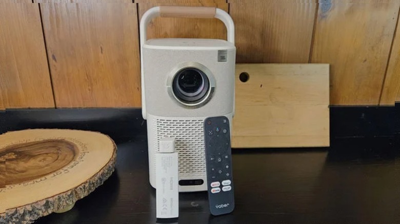 The Yaber T2 Plus Outdoor Projector on a black shelf with a wood background with its remote and dongle resting on the front.