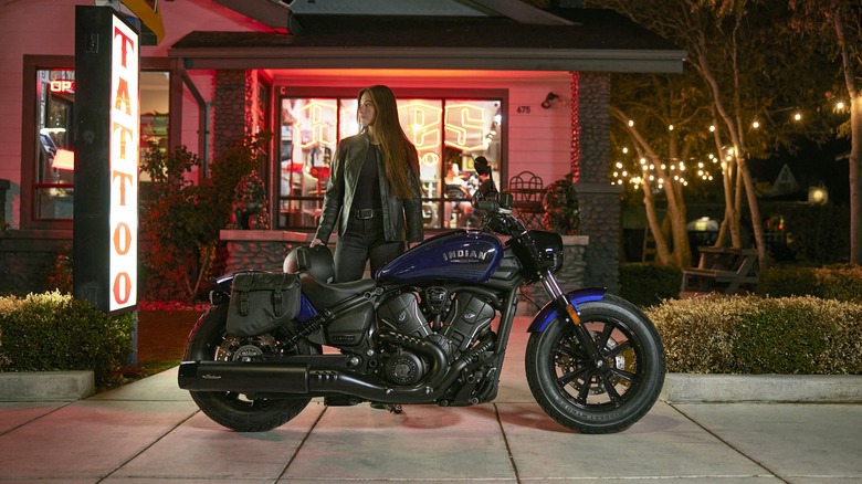 A woman in a leather jacket holding a helmet stands behind an Indian Scout motorcycle parked in front of a tattoo parlour.