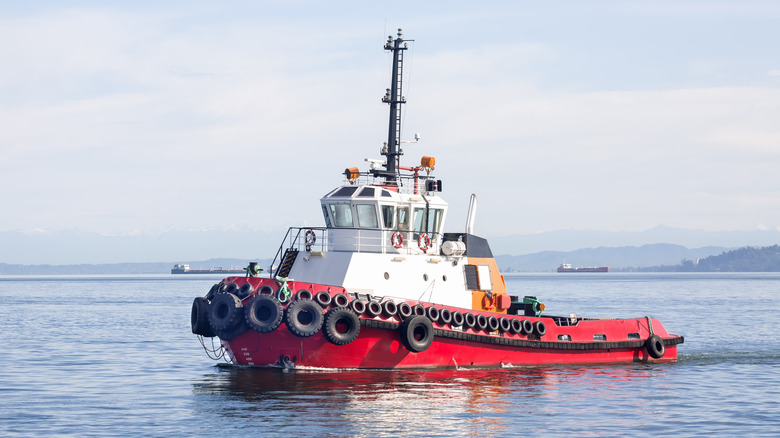 A red tugboat with a white wheelhouse sailing along the hazy hilly coast in the early morning