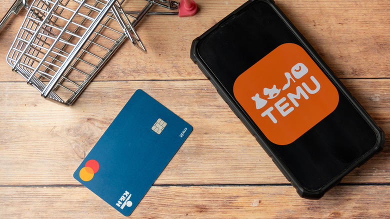Temu logo on a phone screen, a credit card, and a small shopping cart on a wooden surface.
