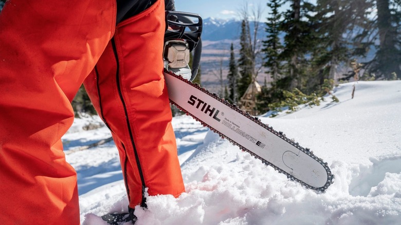 A chainsaw dangles from the hand of a man in orange pants in a winter landscape