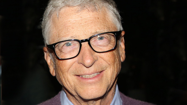 Recent photo of Bill Gates wearing a blue shirt and purple/gray sweater in front of a black background.