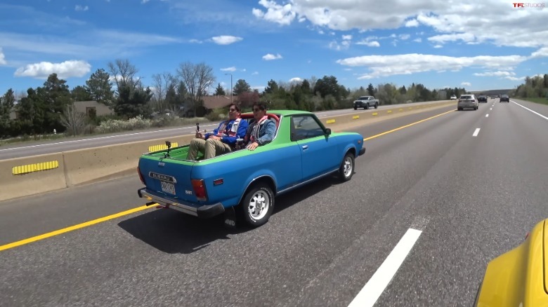 A green and blue first-gen Subaru Brat moving down a freeway with two men in the pickup bed jump seats.